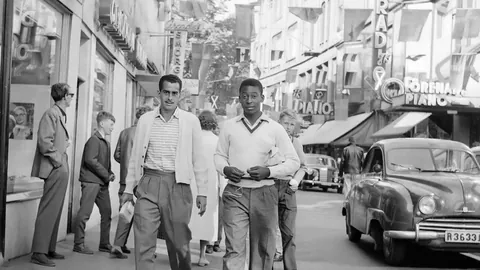 Zito (left) and Pelé walking along a shopping street in Gothenburg during the 1958 World Cup, a small car parked at the kerb.