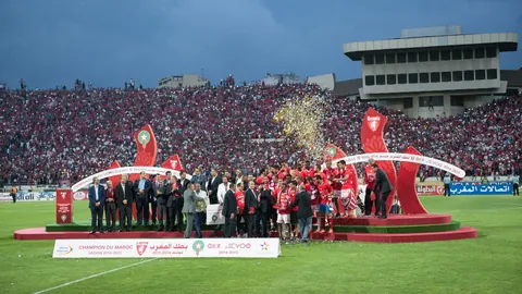 Players in red on a trophy podium in front of a packed stadium, under a Champion du Maroc 2014-2015 banner.